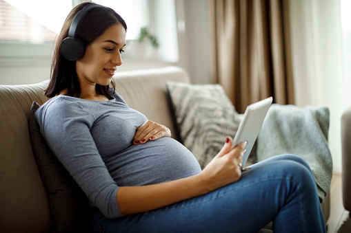 Pregnant woman sitting on couch with tablet and headphones on